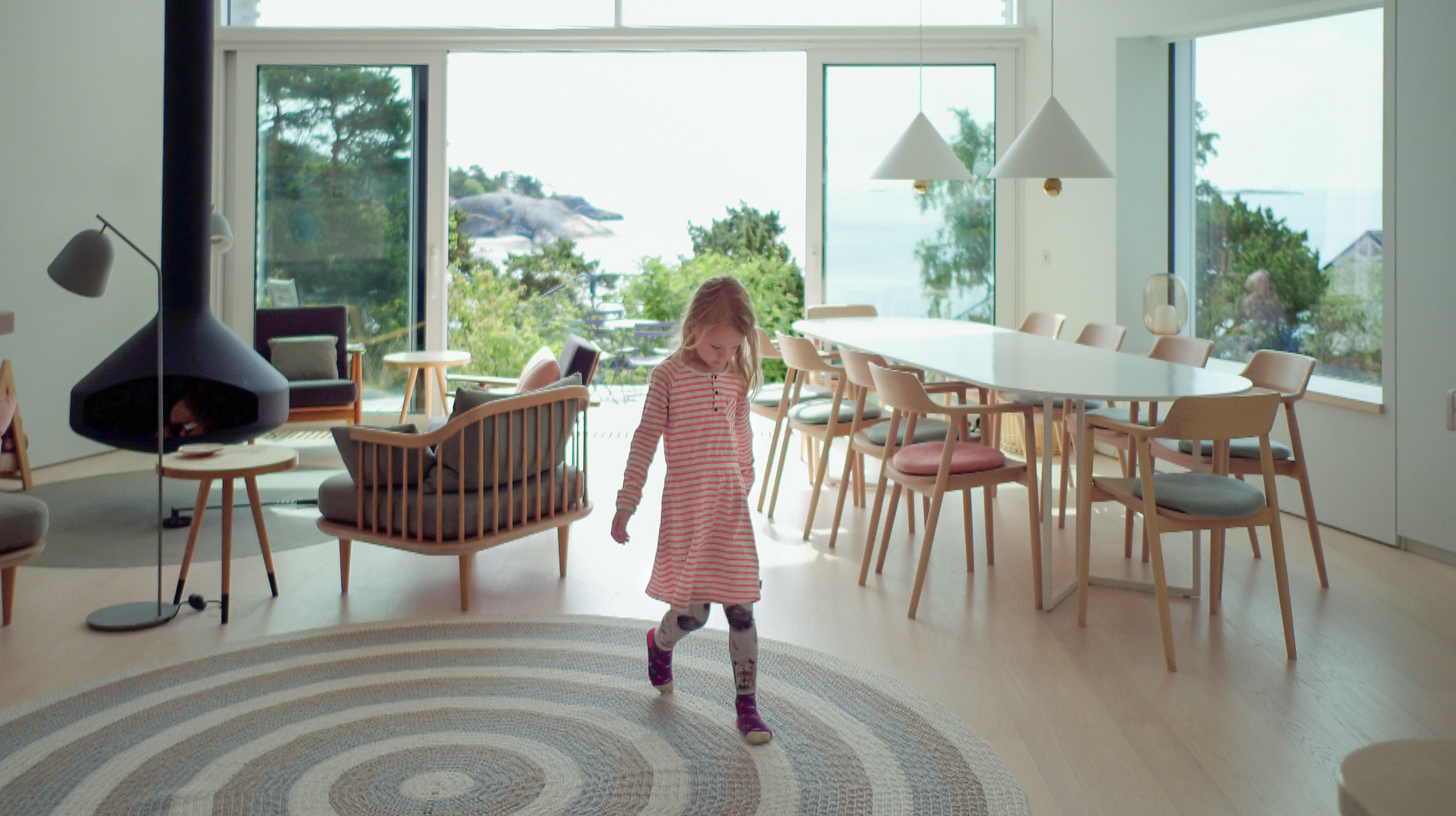 Small girls walking along an pattern of a rug in a room with large windows and sea behind her
