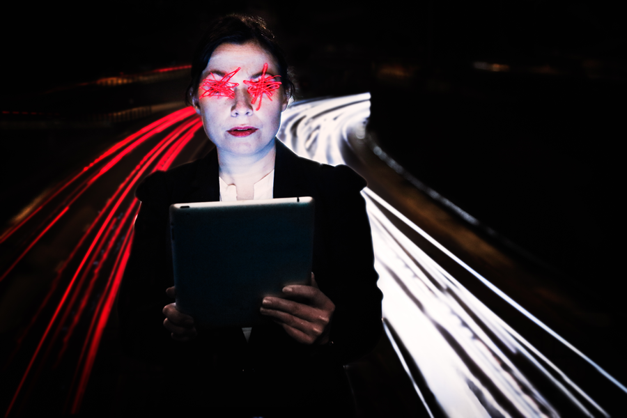 A woman standing in front of a motorway lit by headlights, holding a laptop. Her eyes are smudged out by light trails.